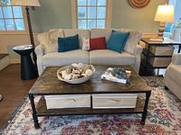 Front view of the industrial style coffee table in a living room setting showing wood top, black metal frame, lower shelf with two beige fabric bins, and metal caster wheels.