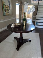 Front and side view of round wooden pedestal table with mahogany finish. Table is set on beige carpet in a foyer with stairs in the background.