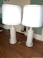 Photo of two matching boho style table lamps with white drum shades on a wooden floor near a door, showing the front and side view.