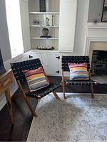 Two modern wooden foldable side chairs with black woven fabric seats and backs photographed with multicolored striped pillows, placed near a fireplace and built-in cabinetry.