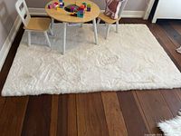 Photo of a 4x6 rectangular white faux fur rug laid on hardwood floor with a small children's table and chairs on top.