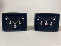 Photo shows two black display boxes each containing a necklace, pendants, and pairs of earrings with clear and pink crystals.