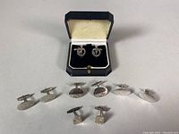 Five pairs of 925 sterling silver cufflinks arranged in front of a black and cream velvet cufflink box.