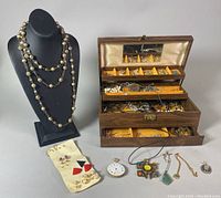 Jewelry box open showing multiple compartments filled with assorted costume jewelry, multiple necklaces and earrings, and some pendants laid out in front of the box and on a black necklace display bust.