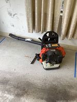 Full side view of the Tanaka backpack leaf blower showing the black nozzle, orange engine housing, and white base tank.