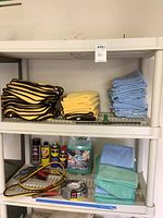 Shelving unit with stacks of yellow and blue microfiber cloths, aerosol spray cans including WD-40, bottle of car wash liquid, jumper cables, tape roll, and other car care supplies