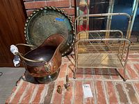 Photo of a large round copper tray with decorative patterning and scalloped edge, a copper bucket with ceramic handles, a small brass milk jug, and a brass lamp with ceramic handle, all placed on red brick floor in front of a fireplace.