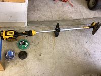 Full view of Dewalt 20V Max XR cordless string trimmer with aluminum shaft, black handle, and attached battery. Two extra spools of trimmer string visible on floor.