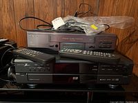 Hitachi FX621 VHS VCR stacked on top of Zenith DVC2515 DVD player, both with respective remotes on top and power cords visible, placed against a wood-paneled background.