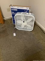 Front view of Lasko 20 inch box fan with white plastic housing and original box behind it on floor mat.