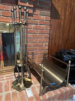 Photo showing the full fireplace tool set with stand next to the log rack, all brass tone metal, placed beside a brick fireplace.