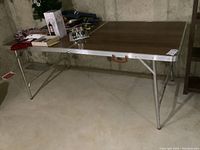 Full view of the rectangular folding table with metal frame and wood grain top with scratches. Legs extended and table set on concrete floor.