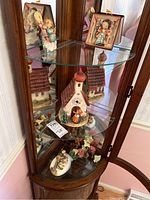 Photo showing wooden corner cabinet with glass shelves housing the clock building sculpture and small figurines around it.