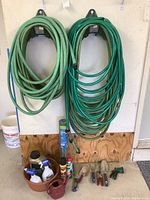 Photo of multiple green garden hoses hung on wall brackets with assorted garden sprays and tools below them.