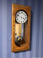 Full view of the Howard Miller wooden wall clock hanging on a blue striped wallpaper wall, showing the white clock face, pendulum, and brass details.