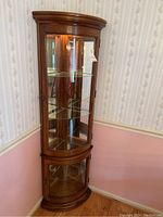Front view of the wooden corner display cabinet showing curved glass doors, light inside, and overall tall structure.