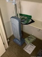 Light blue Oreck XL upright vacuum cleaner standing upright near shelf with plastic sealed vacuum bags and storage bags on the floor next to it.