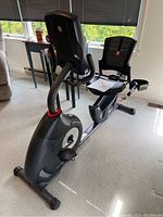 Side and angle view showing black Schwinn recumbent bike with ergonomic seat, backrest, foot pedals, and digital display console, plus the manual on the seat.