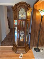 Full view of the oak-finished tall grandfather clock with glass panels on front and sides, showing weights and pendulum inside, positioned near a floor lamp.