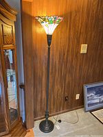 Full view of the floor lamp on, showing stained glass shade emit colorful light, and metal base.