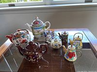 Photo of eight assorted ceramic and metal tea pots on a glass table near a window.