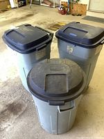 Front view of three Rubbermaid Roughneck trash cans with lids; two have wheels, all are heavily soiled and weathered.