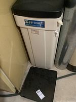 Zerosoft branded white plastic water conditioning tank with black lid placed on floor near wall and pipe. A small spout is visible on the side.