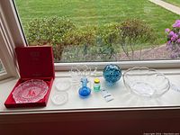 Overview of all items on windowsill: Waterford boxed dish, Val Saint Lambert bowl, two glass coasters, blue glass pitcher, mini jar, art glass hanging orb, black seal figure.