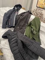 Four ladies large coats and jackets on a bed, including a navy Helly Hansen sailing jacket, a black coat, a green quilted jacket, and a black quilted jacket.