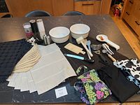 Overview photo of lot items on a kitchen counter including Corningware bowls, place mats, aprons, and kitchen utensils.