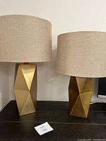 Two gold-tone metal base lamps with faceted geometric design and beige tweed lampshades side by side on black surface