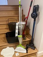 Smart Living Steam Mop in white and lime green plastic standing on wooden stairs with mop pads and charger, next to cordless vacuum and red Bee Mop, two brown bins and black basket behind