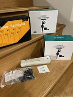 Photo of all spotlight boxes arranged on stairs showing 8-pack Meikee box in black and orange next to two white and teal Edishine boxes and installation parts.