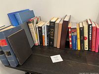 Photo showing 24 various books lined up on a dark surface, spines visible with titles covering diverse subjects and genres.