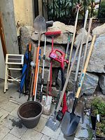 Photo showing the collection of garden tools including shovels, broom, cordless trimmer, and plant pots against a stone wall.