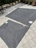 View of two rectangular outdoor rugs lying flat on a light stone patio, showing the lighter side of the woven texture. Larger rug approximately 6'x9', smaller rug approximately 4'x6'.