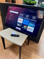 Front full view of the Sanyo 32" smart TV turned on with Roku TV interface visible on screen, with remote control on nearby stool.