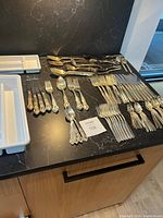 Overview of the full assortment of silverware laid out on a black countertop showing various utensils including forks, spoons, and knives with ornate handles.
