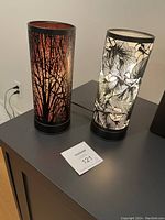 Front view showing two cylindrical touch lamps with distinct nature-themed shades; one with tree branches and orange glow, the other with white floral design.