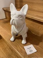 Front view of the white ceramic French Bulldog figurine sitting on wooden stairs with a small pendant around its neck.