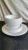 Stack of 11 white ceramic bowls with green rim on top of stacked plates