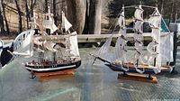 Two wooden sailboat models on a glass table outdoors. The model on the left has black and white paint with multiple white sails and detailed rigging. The model on the right has a blue hull and white sails.