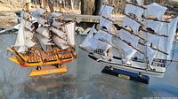 Two model sailboats on a glass table outdoors with trees in the background. One is HMS Victory and the other is Whaling Ship Clipper 1846.