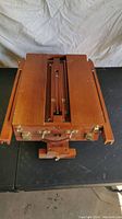 Folded collapsible wooden easel resembling a briefcase with brass clasps and handle, showing the easel compacted for transport/storage.