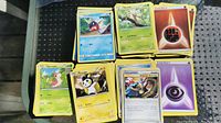 Multiple stacks of Pokémon trading cards showing various Pokémon and energy cards on top of each stack on a green outdoor table.