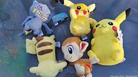Five Pokémon plush toys laid out on a blanket including Pikachu, Chimchar, Psyduck, Croagunk, and a striped beige Pokémon plush.