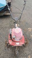 Front view of red rototiller showing handlebars, engine cover, and wheels on gravel ground.