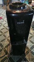 Front view of Primo black water dispenser showing circular water bottle holder on top and drip tray at bottom.