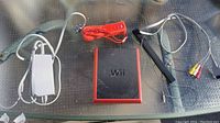Top down photo of Nintendo Wii console (black and red), white power adapter, red Wii remote with wrist strap, white sensor bar, and AV composite cables on a glass table.