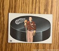 Front view of 1962-63 Parkhurst Sid Abel hockey card showing player standing on a large hockey puck with Detroit Red Wings logo, wearing team jacket and gloves, holding hockey stick. Card number 34 visible.